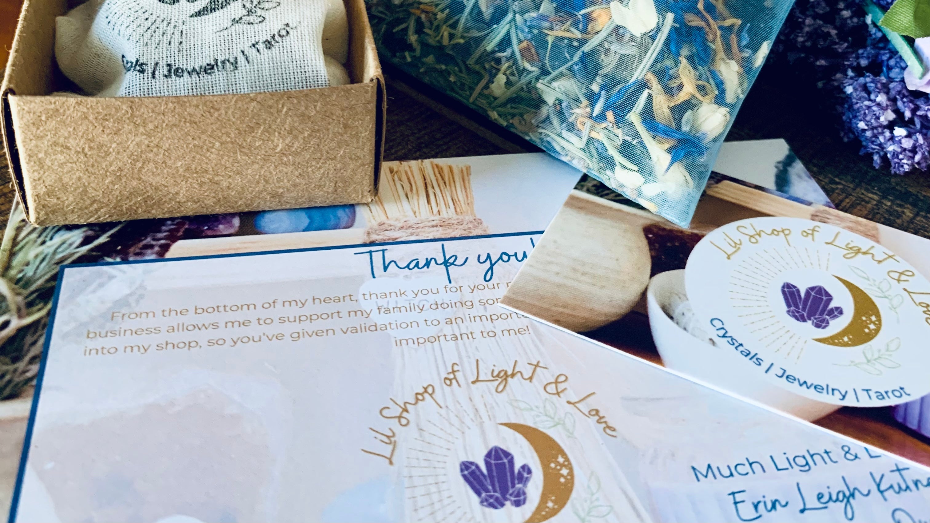 Lil shop of light & love packaging, including business card, thank you card, herb & crystal mix in a small sachet, a small craft jewelry box with a cloth bag inside, stamped with the lil shop of light & love logo of a crescent moon, crystals, & vines