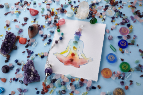 watercolor painting of outline of meditating person with crystals representing each chakra and matching watercolor rainbow in the outline. crystals are scattered around on a light blue surface.