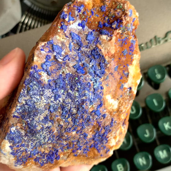 A raw chunk of azurite with malachite & mica being turned to see all the angles in front of a green typewriter.