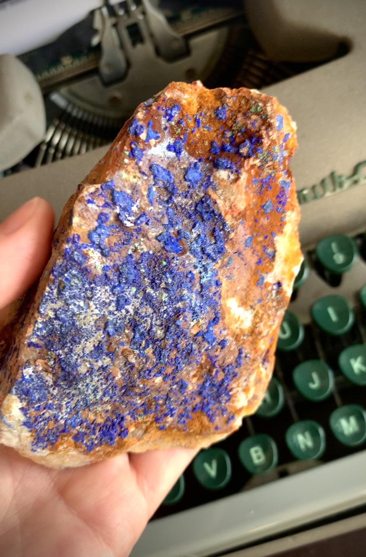 A raw chunk of azurite with malachite & mica being turned to see all the angles in front of a green typewriter.