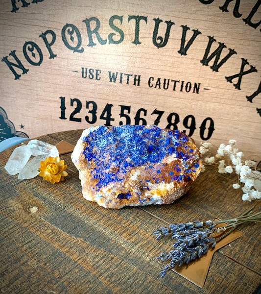 A raw chunk of azurite with malachite & mica on a wooden surface with flowers and quartz crystals around it. A ouija board is the background.
