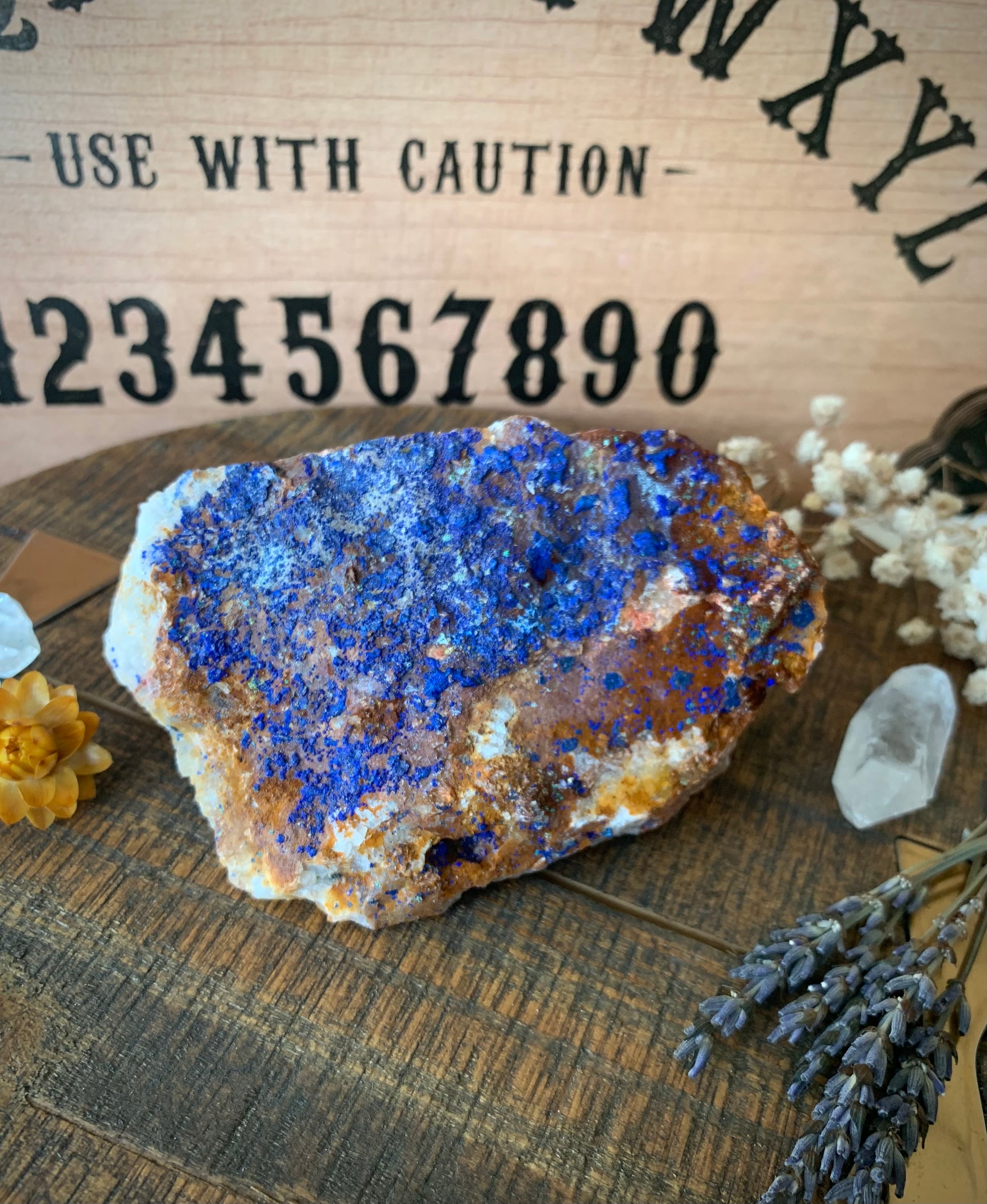 A raw chunk of azurite with malachite & mica on a wooden surface with flowers and quartz crystals around it. A ouija board is the background.