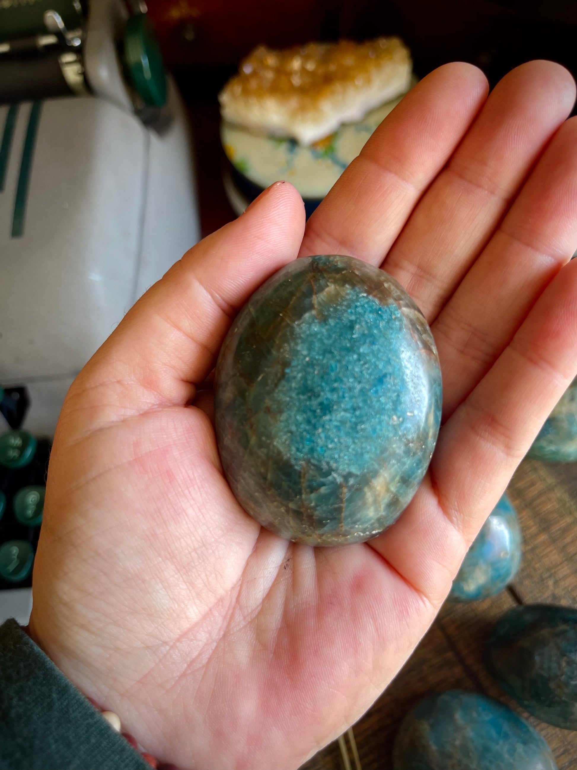 Blue apatite palm stone held in a hand In front of a desk with pretty things & a typewriter 