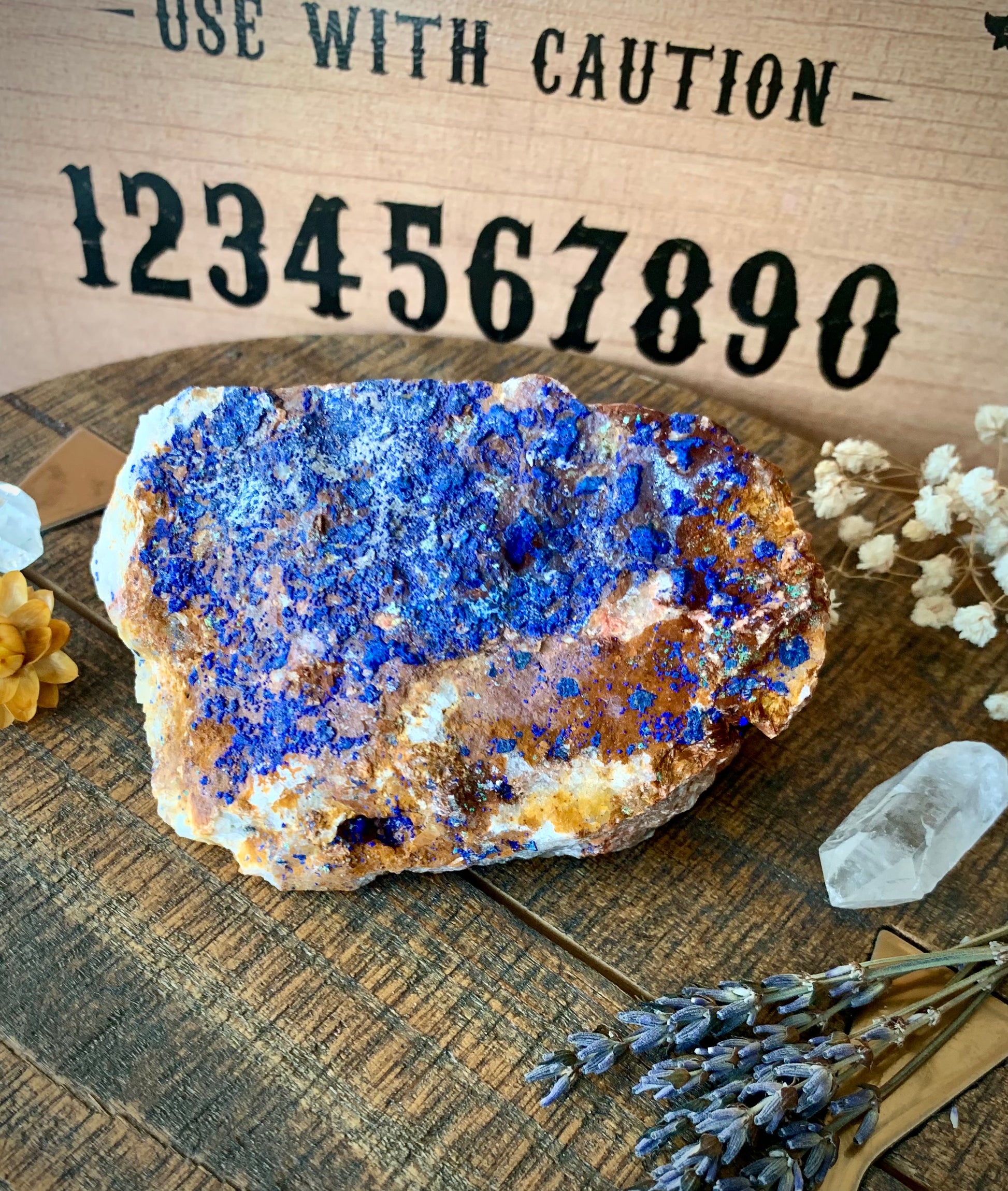 A raw chunk of azurite with malachite & mica on a wooden surface with flowers and quartz crystals around it. A ouija board is the background.